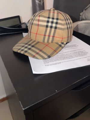 Rutig Burberry keps i beige - Säljer en klassisk Burberry keps i beige med det ikoniska rutiga mönstret i svart, vitt och rött. Justerbar baktill med svart spänne och Burberry London England-logga. Perfekt accessoar för dig som vill sticka ut med stil. Den har dock en skada på knappen där back på kepsen samt lite blått färg på den som man tydligt ser på bild! Vid frågor är d bara att ställa dem tack!
