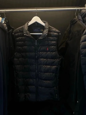 Svart dunväst från Polo Ralph Lauren - Snygg svart dunväst från Polo Ralph Lauren med quiltad design och dragkedja framtill. Västen har ståkrage och den klassiska röda loggan broderad på bröstet. Perfekt att slänga över en hoodie eller tröja för extra värme och stil.