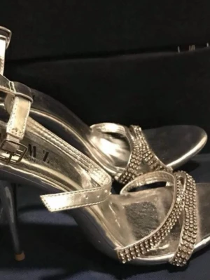 Silvriga sandaler med strass och klack - Snygga silvriga sandaler med smala remmar dekorerade med glittrande strass. Skorna har öppen tå, hög klack och justerbart spänne vid ankeln. Perfekta för dig som vill sticka ut med lite extra bling.passar st/38-39