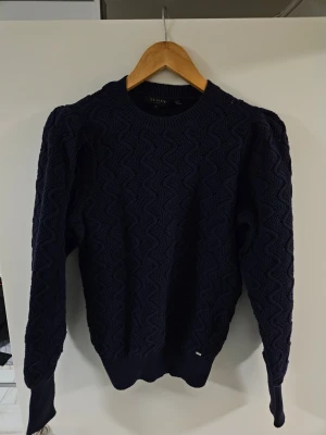 Mörkblå stickad tröja Ted Baker - Snygg mörkblå stickad tröja från Ted Baker med vågigt mönster över hela plagget. Tröjan har rund halsringning, långa ärmar och ribbade muddar vid ärmslut och nederkant. Perfekt för dig som gillar stilrena och klassiska plagg.