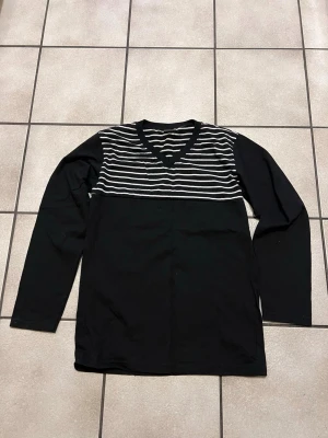 Svart långärmad tröja med ränder 100% bomull  - Snygg svart långärmad tröja med vita horisontella ränder över bröstet och V-ringad hals. Tröjan har en enkel och stilren design som passar till det mesta. Tillverkad i mjukt bomullstyg för skön känsla.