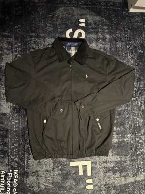 Svart vintage jacka från Polo Ralph Lauren - Klassisk svart jacka från Polo Ralph Lauren med dragkedja framtill, krage i manchester och broderad logga på bröstet. Jackan har långa ärmar, knappdetaljer vid ärmslut och två fickor med knappstängning. Perfekt för en stilren look.