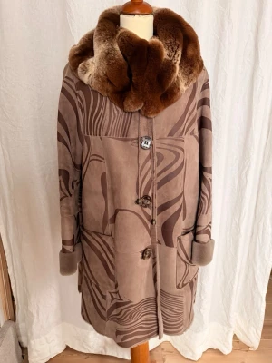 Brun vintage pälsjacka med mönster - Unik brun pälsjacka med bred, fluffig krage i äkta päls och abstrakt mönster i olika bruna nyanser. Jackan har stora fickor, dekorativa knappar och uppvikta ärmslut. Perfekt för dig som vill sticka ut med en retrolook.