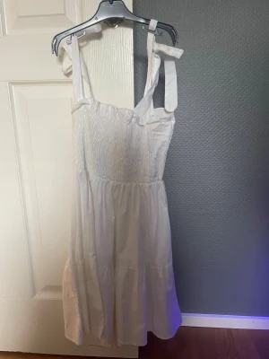 Vit sommarklänning från RYVL - Söt vit klänning från RYVL med smock upptill och breda axelband som knyts på axlarna. Klänningen har en luftig, volangig kjol och är perfekt för varma dagar. Tillverkad i mjuk bomull som känns skön mot huden.