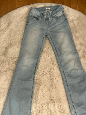 Ljusblå bootcut jeans från Nelly med broderi - Säljer ett par ljusblå bootcut jeans med snyggt broderi i rosa på bakfickorna. Jeansen har låg midja och klassisk femficksdesign. Perfekta för dig som gillar en avslappnad men trendig look.