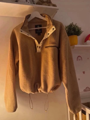Beige fleece-tröja  - Mysig beige fleece-tröja  copmmed halvknäppning framtill, bröstficka med dragkedja och dragsko i midjan. Tröjan har hög krage och långa ärmar, perfekt för kalla dagar. Materialet är mjukt och fluffigt för extra komfort.
