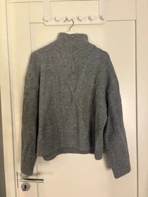 Grå stickad tröja med halvzip -  grå stickad tröja med hög krage och halvzip framtill. Tröjan har ett diskret kabelstickat mönster, ribbade muddar och avslappnad passform. Perfekt för lager på lager och chill dagar. Passar bra för alla årstider!