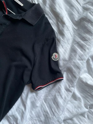 Svart Moncler pikétröja  - Säljer en fet Moncler piké i storlek M.                 Pris kan såklart diskuteras.                                Tröjan har krage, knappar framtill och Moncler-logga på vänster ärm. Perfekt för en stilren och sportig look.