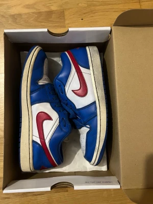 Nike Air Jordan 1 Low blå/röd/vit - Säljer ett par Nike Air Jordan 1 Low sneakers i blått, vitt och rött. Skorna har klassisk låg siluett, perforerad tåbox och den ikoniska röda swoosh-loggan på sidan. Ovandelen är i läder och de har blå snören samt vit mellansula. Kvitto och box kommer med.