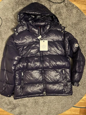 Mörkblå dunjacka från Moncler - Snygg mörkblå pufferjacka från Moncler med huva och dragkedja framtill. Jackan har två framfickor med dragkedja och klassisk Moncler-logga på ärmen. Insidan har ett unikt serietidningsmönster. Perfekt för kalla dagar. NFC tag fungerar