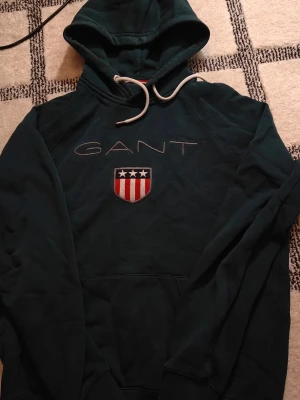 Mörkgrön hoodie från GANT - Snygg mörkgrön hoodie från GANT med broderad logga och amerikanskinspirerat sköldmärke på bröstet. Hoodien har vita dragsnören och en klassisk känguruficka framtill. Perfekt för dig som gillar stilrena och sportiga plagg.