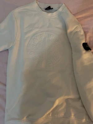 Vit sweatshirt från Stone Island - Vit sweatshirt från Stone Island med stort präglat logotyptryck på bröstet och den klassiska svarta patchen med gulgrön kompass på ärmen. Tröjan har rund hals och ribbade muddar. Perfekt för en clean och stilren look.
