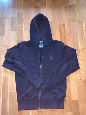Mörkblå zip hoodie från Gant - Snygg mörkblå hoodie från Gant med dragkedja framtill och klassisk huva med dragsko. Broderad Gant-logga på bröstet och två fickor fram. Perfekt för en avslappnad och stilren look. Tillverkad i mjukt bomullsmaterial. Vid frågor är det bara att höra av sig!