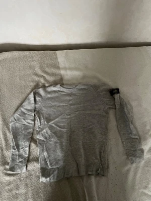 Grå stickad tröja Stone Island Junior - Snygg grå stickad tröja från Stone Island Junior med klassisk rund hals och ribbade muddar. Tröjan har den ikoniska Stone Island-loggan på vänster ärm. Perfekt för dig som gillar stilrena och trendiga plagg. Barnstorlek 14/170 men sitter lowkey som en S. Vet ej om den är äkta eller inte 🫃🤷‍♂️
