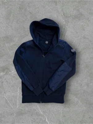 Mörkblå hoodie från Colmar - Snygg mörkblå hoodie från Colmar med dragkedja framtill och en klassisk huva. Ärmarna har ett diskret Colmar-märke på vänster sida. Hoodien är tillverkad i mjukt material och har ribbade muddar vid ärmslut och nederkant. Perfekt för en avslappnad och stilren look.