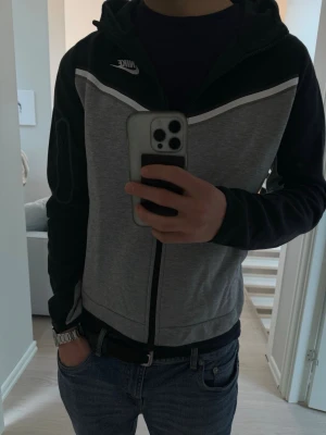 Nike tech fleece - Snygg Nike tech fleece i grått och svart med vitt märke. Tröjan har dragkedja , två fickor och en huva. Tröjan har en extra ficka på ärmen. Perfekt för en sportig eller avslappnad stil, STORLEK 158-170 cm men passar som en vanlig S.