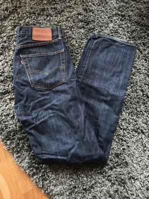 Levi's 501 mörkblå jeans straight fit - Klassiska Levi's 501 jeans i mörkblå tvätt med raka ben och normal passform. Jeansen har fem fickor, synliga kontrastsömmar och den ikoniska läderpatchen bak i midjan. Tillverkade i slitstark bomullsdenim och passar perfekt till en avslappnad stil.