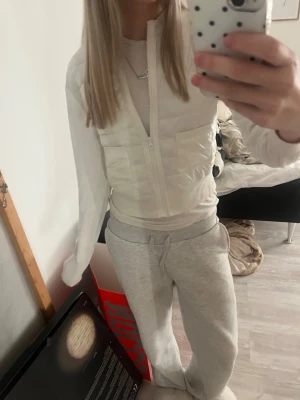 Vit dunväst med dragkedja - Säljer en stilren vit dunväst med dragkedja framtill och två fickor. Västen har en kort, boxig passform och quiltad design som ger en modern look. Perfekt att slänga över en hoodie eller tröja för extra värme. Enkel att matcha med olika outfits.