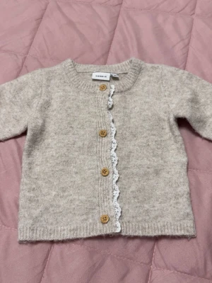 Beige kofta med träknappar & spets - Supersöt beige kofta från Name it med rund halsringning, träknappar och vit spetsdetalj längs knäppningen. Koftan är stickad i mjukt material och har långa ärmar. Perfekt för en mysig och stilren look.