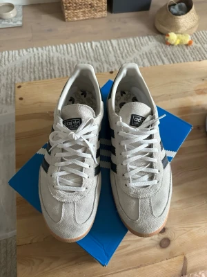Adidas spezial i ljus mocka - Snygga Adidas Samba sneakers i ljusgrå mocka med klassiska svarta ränder på sidorna och gummisula. Skorna har rund tå, snörning och retrovibbar. Perfekta för dig som gillar stilrena och tidlösa sneakers.