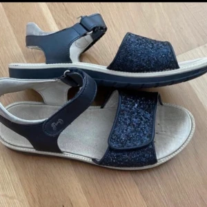 Marinblå sandaler med glitterdetaljer storlek 39 - Snygga svarta sandaler med glittriga band över foten och justerbar rem med kardborre runt ankeln. Sandalerna har platt sula och öppen tå, vilket ger en bekväm och luftig känsla. Perfekta för varma dagar och passar till många olika outfits. Passar både barn och vuxen. 