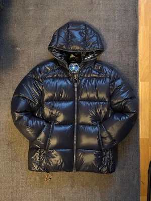 Svart dunjacka från Save The Duck - Snygg svart dunjacka från Save The Duck i modellen Edgar. Jackan har en glansig finish och padded design, färgen ”blue black”. Jackan har huva, dragkedja framtill och två sidofickor. Perfekt för kalla dagar och ger en cool, sportig look. Storlek: M