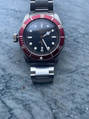 Tudor Black Bay klocka röd bezel - Snygg Tudor Black Bay dykarklocka med röd bezel och svart urtavla. Klockan har tydliga index, stora visare och boett samt länk i rostfritt stål. Perfekt accessoar för dig som gillar klassisk och sportig stil.