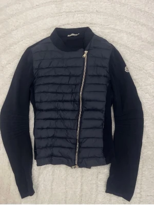 Svart cardigan jacka från Moncler - Snygg svart cardigan jacka från Moncler med quiltad framsida och asymmetrisk dragkedja. Ärmarna är stickade och har Moncler-logga på ena armen. Perfekt för lager på lager och en stilren look. Dragkedjan har gått sönder lite