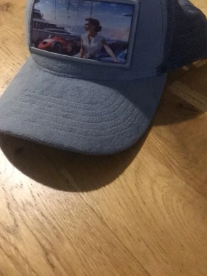 Grå truckerkeps med bildpatch - Snygg grå truckerkeps med mesh på sidorna och en stor patch framtill med motiv av en kvinna och en röd sportbil vid en hamn. Kepsen har böjd skärm och justerbar baksida. Perfekt för dig som gillar unika accessoarer.