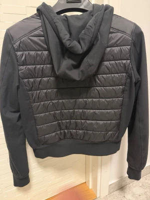 Svart pufferjacka från Parajumpers - Snygg svart pufferjacka från Parajumpers med quiltad front och rygg, huva och dragkedja framtill. Jackan har ribbade muddar och två fickor med dragkedja. Perfekt för dig som vill ha en sportig och modern look.