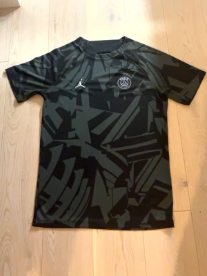 Svart PSG x Jordan t-shirt - Säljer en svart t-shirt från PSG x Jordan med coolt abstrakt mönster i mörkgrått. T-shirten har korta ärmar, rund halsringning och PSG-logga samt Jumpman-logga på bröstet. Tillverkad i ett lätt och skönt syntetmaterial som passar perfekt till sport och streetwear.