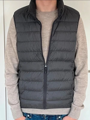 Mörkgrå dunväst med dragkedja - Snygg mörkgrå dunväst med hög krage och horisontella sömmar. Västen har en stilren look med dragkedja framtill och är lätt vadderad för extra värme. Perfekt att slänga över en hoodie eller tröja för en chill och trendig stil. Uniqlo