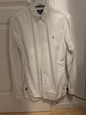 Vit skjorta från Polo Ralph Lauren - Klassisk vit skjorta från Polo Ralph Lauren med broderad logga på bröstet. Skjortan har lång ärm, knappar framtill och en stilren krage. Perfekt för en clean och tidlös look.