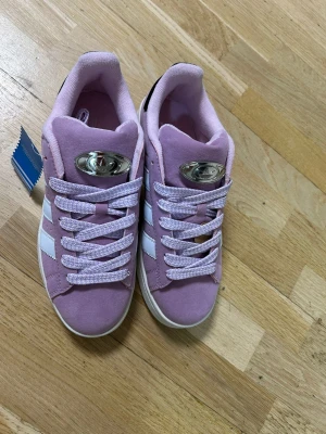 Adidas Campus rosa sneakers mock pipeline - Snygga Adidas Campus sneakers i ljusrosa mocka med vita klassiska ränder på sidorna och svart detalj vid hälen. Skorna har rund tå, platt sula och snörning med matchande rosa/vita skosnören. Metallmärke med Adidas-logga på plösen.