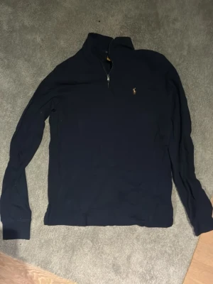Mörkblå halvzip från Polo Ralph Lauren - Mörkblå långärmad tröja från Polo Ralph Lauren med halv dragkedja och klassisk broderad logga på bröstet. Tröjan har hög krage och är tillverkad i mjukt material, perfekt för lager-på-lager-stil.