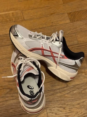 Vita Asics sneaker - Fräscha sneakers från Asics i vitt meshmaterial med röda och silverfärgade detaljer. Skorna har snörning, platt sula och svart vadderad insida för extra komfort. Perfekta för en sportig och trendig look.