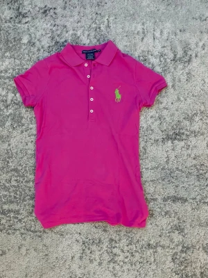 Rosa pikétröja från Ralph Lauren - Säljer en rosa pikétröja från Ralph Lauren med klassisk krage och knappar framtill. Tröjan har korta ärmar och den ikoniska gröna logotypen broderad på bröstet. Perfekt till jeans eller kjol för en sportig och stilren look.