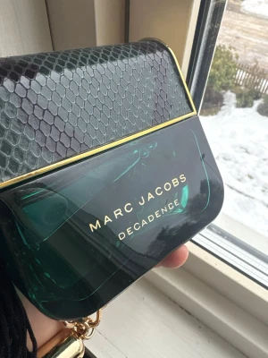 Marc Jacobs Decadence Eau de Parfum - Marc Jacobs Decadence Eau de Parfum i en lyxig smaragdgrön flaska med gulddetaljer och lock i ormmönstrad design. Flaskan har en unik form som påminner om en handväska och kedja i guld. Doften är elegant och modern.