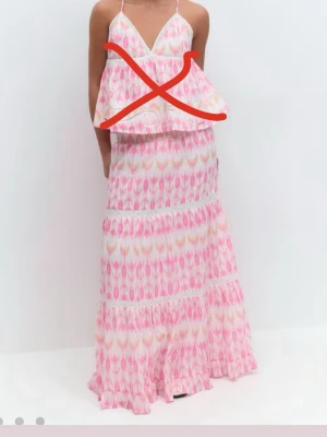 Långkjol i rosa mönster med volanger - Säljer en långkjol med volanger i ett färgglatt rosa, vitt och gult mönster. Kjolen har smock i midjan och spetsdetaljer mellan volangerna. Perfekt för dig som gillar boho-vibbar och vill ha en luftig kjol till sommaren.