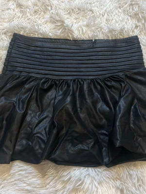 Svart kort kjol i skinnimitation - Snygg svart kort kjol i skinnimitation med bred, ribbad midja och lätt utställd form. Perfekt för dig som vill ha en edgy och trendig look. Kjolen har en glansig finish och markerad midja som ger en cool siluett. Har tagit av lappen för den skav men har för mig den är från ginatricot eller HM!