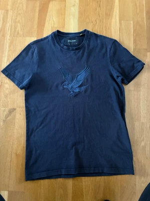 Mörkblå t-shirt från Lyle & Scott - Snygg mörkblå t-shirt från Lyle & Scott med broderad fågel på bröstet. Klassisk rund halsringning och korta ärmar. Tillverkad i mjuk bomull som känns skön mot huden. Perfekt för dig som gillar stilrena plagg med en twist.