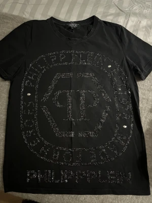 Svart Philipp Plein t-shirt med paljetter - Snygg svart t-shirt från Philipp Plein med stort logotryck och text framtill, dekorerad med glittrande paljetter. Klassisk rund halsringning och korta ärmar. Perfekt för dig som vill sticka ut med en exklusiv vibe.