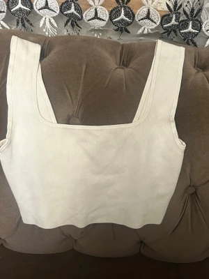 Vit ribbad croppad linnetopp - Säljer en vit croppad linnetopp med breda axelband och fyrkantig urringning. Toppen är ribbad och har en enkel, stilren design som passar perfekt till jeans eller kjol. Perfekt för dig som gillar minimalistisk stil.