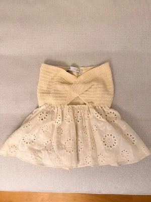 Beige axelbandslös topp från Zara - Söt beige axelbandslös topp från Zara med virkad överdel och broderad, utsvängd nederdel i bomull. Toppen har fina spetsdetaljer och ett omlottmönster upptill. Perfekt för varma dagar och sommarens alla tillfällen.