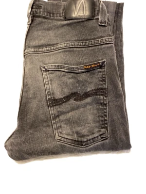 Grå jeans från Nudie Jeans - Snygga grå jeans från Nudie Jeans med klassisk femficksmodell och svarta broderade detaljer på bakfickorna. Jeansen har en rak passform och är tillverkade i mjukt denimtyg. Perfekta för en avslappnad och trendig look.Skick 6/10