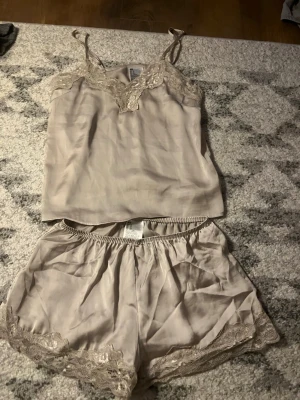 Beige satinset med spetsdetaljer - Snyggt tvådelat set i beige satin med linne och shorts. Linnet har smala axelband och spets vid halsringningen, medan shortsen har elastisk midja och spets längs bensluten. Perfekt för en mjuk och lyxig känsla hemma. Är från h&m