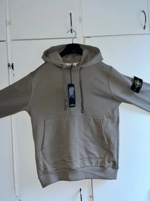 Beige hoodie från Stone Island - Snygg beige hoodie från Stone Island med klassisk patch på vänster ärm. Hoodien har dragsko i huvan och en stor magficka framtill. Tillverkad i mjukt bomullsmaterial och har ribbade muddar vid ärmslut och nederkant. Perfekt för en avslappnad streetwear-look.