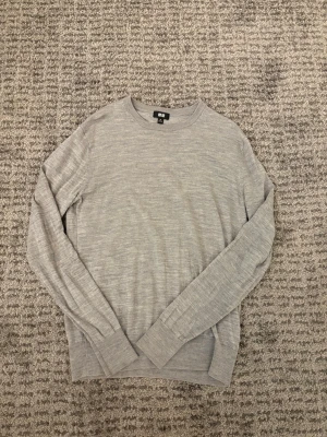 Ljusgrå stickad tröja från Uniqlo - En stilren ljusgrå stickad tr sop från Uniqlo med rund halsringning och långa ärmar. Tröjan har ribbade muddar vid ärmslut och nederkant. Perfekt för lager på lager och enkel att matcha med olika outfits.