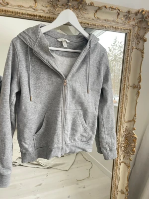 Grå zip hoodie från H&M - Klassisk grå hoodie från H&M med dragkedja framtill, huva med dragsko och två stora fickor. Tillverkad i mjuk bomullsblandning som är perfekt för chill dagar. Ribbade muddar vid ärmslut och nederkant ger en snygg passform.