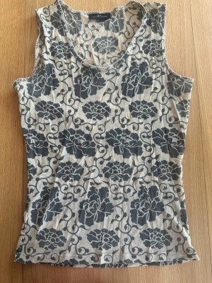 Blommig ärmlös meshtopp - Snygg ärmlös linnetopp från Day med gråblått blommönster på beige botten. Toppen har rund halsringning och är i ett mjukt, tunt material som känns skönt mot huden. Perfekt för dig som gillar en chill och trendig stil.